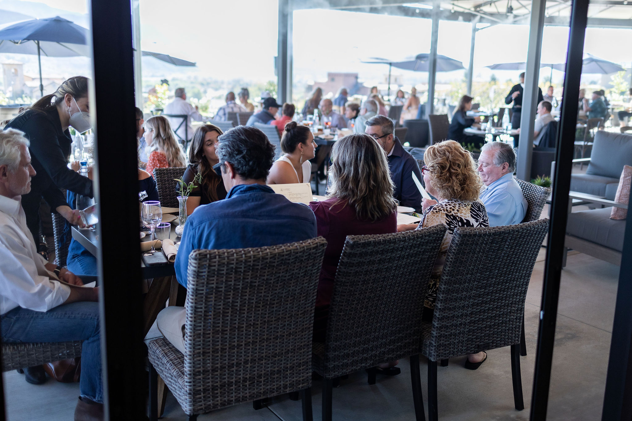 A group of people dining at a restaurant with outdoor seating. There are multiple tables with diners, and servers in the background. The setting features umbrellas and a scenic view.