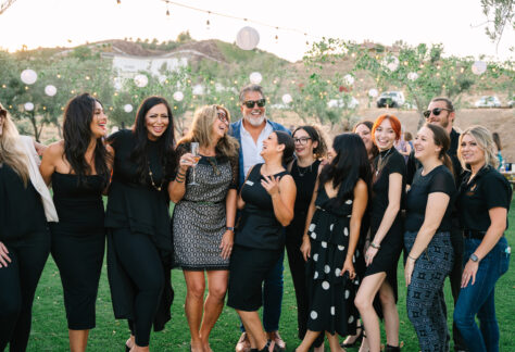 A group of people smiling and posing together at an outdoor event in a garden setting. They are dressed in various styles, with a mix of black outfits, and the background features greenery and decorative lights.