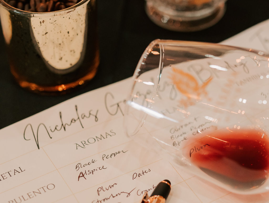 A wine tasting sheet featuring the name "Nicholas Gregory" with aromas listed, alongside a tipped wine glass spilling red wine and scattered writing utensils on a dark table.