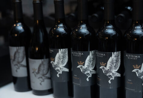 A row of wine bottles from Altísima Winery, featuring dark glass and distinctive labels with an eagle design. The bottles display different vintages, creating a uniform presentation.