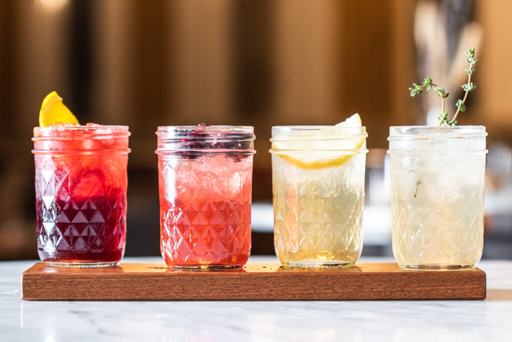 Four mason jars filled with colorful beverages displayed on a wooden tray. The drinks include vibrant red, pink, and clear options garnished with fruit slices and herbs.