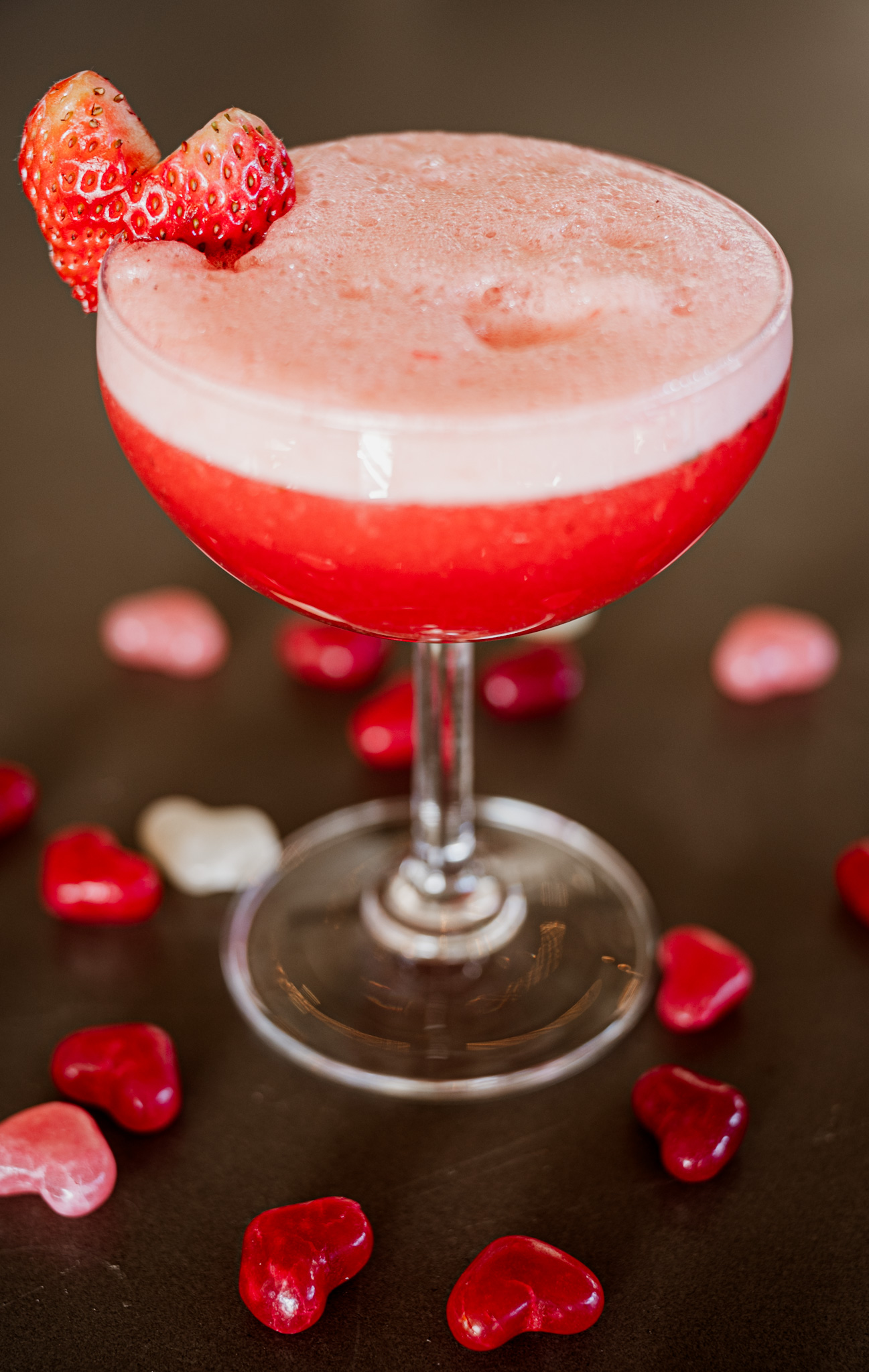 A strawberry cocktail in a coupe glass topped with sliced strawberries, surrounded by colorful heart-shaped candy on a dark surface. The drink features a gradient of pink and red colors.