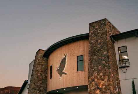 An exterior view of a contemporary building featuring a stone facade and wooden accents, with an eagle emblem prominently displayed above the entrance, set against a dusk sky.