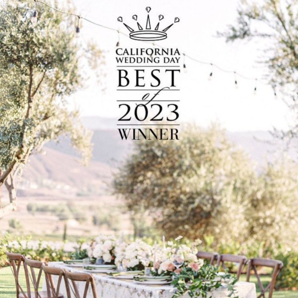 An outdoor wedding reception setup at a scenic location, featuring a long table adorned with floral arrangements. A sign at the top indicates "California Wedding Day BEST of 2023 WINNER." The setting includes trees and hills in the background, ideal for a Temecula wedding venue.