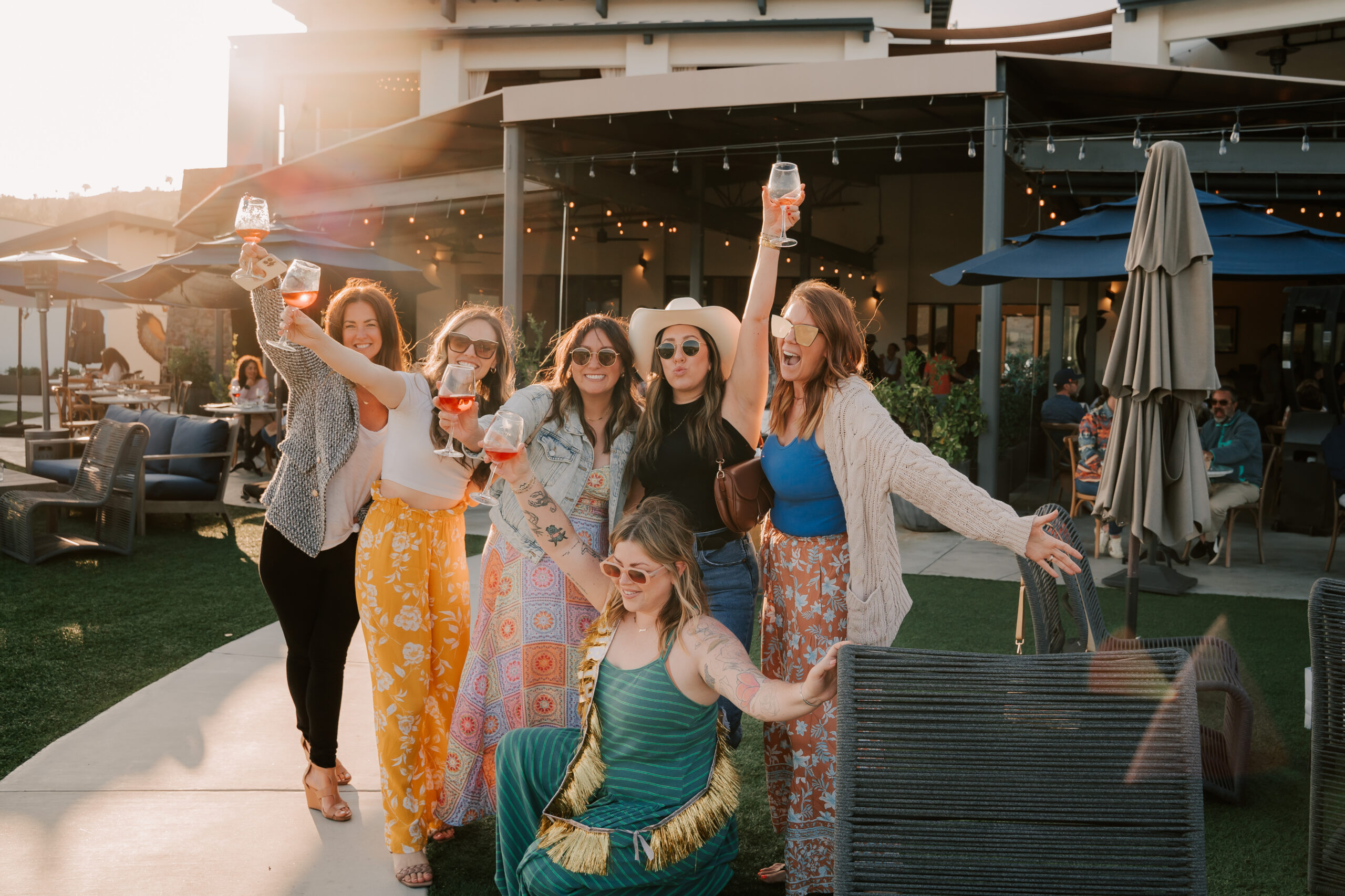 A group of six women celebrating outdoors in a well-lit setting, holding drinks and smiling. They are wearing colorful outfits, surrounded by patio furniture and festive lighting.