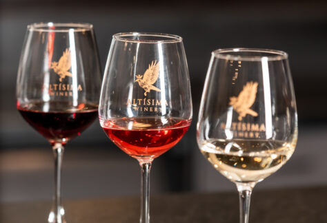 Three wine glasses on a dark countertop, each with a different type of wine: a red wine in a glass on the left, a rosé in the center, and a white wine on the right, all branded with the logo of Altisima Winery.