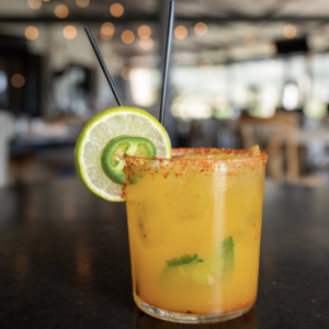 A colorful cocktail garnished with a lime and jalapeño slice, served in a glass with a rim coated in chili powder. Two black straws protrude from the drink, set against a blurred restaurant background.