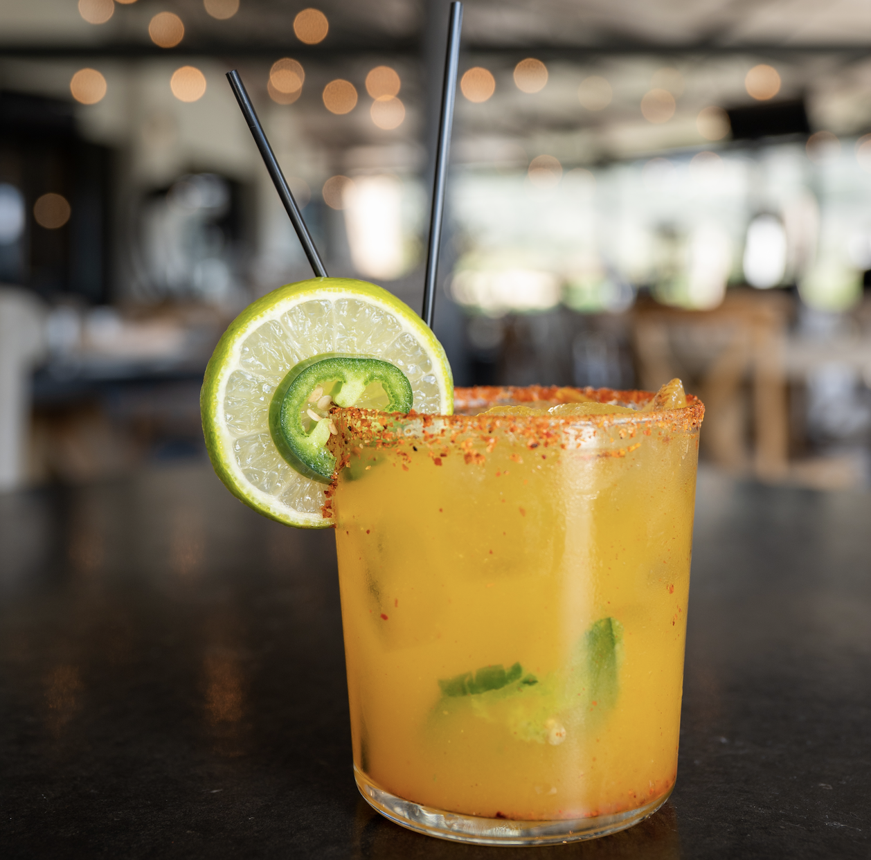 A colorful cocktail garnished with a lime and jalapeño slice, served in a glass with a rim coated in chili powder. Two black straws protrude from the drink, set against a blurred restaurant background.
