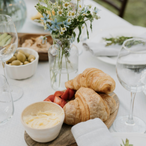 A table setting featuring two golden croissants and sliced strawberries on a wooden platter. A small bowl of butter and a folded napkin are nearby. A glass of water and a vase with assorted flowers add to the rustic dining atmosphere.