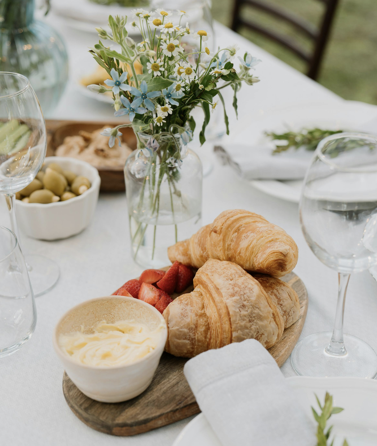 A table setting featuring two golden croissants and sliced strawberries on a wooden platter. A small bowl of butter and a folded napkin are nearby. A glass of water and a vase with assorted flowers add to the rustic dining atmosphere.