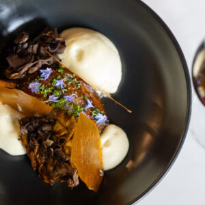 A black bowl contains a serving of mashed potatoes paired with dark mushrooms and garnished with orange crispy elements and small edible flowers. Next to the bowl, there is a glass of red wine resting on a white marble surface.