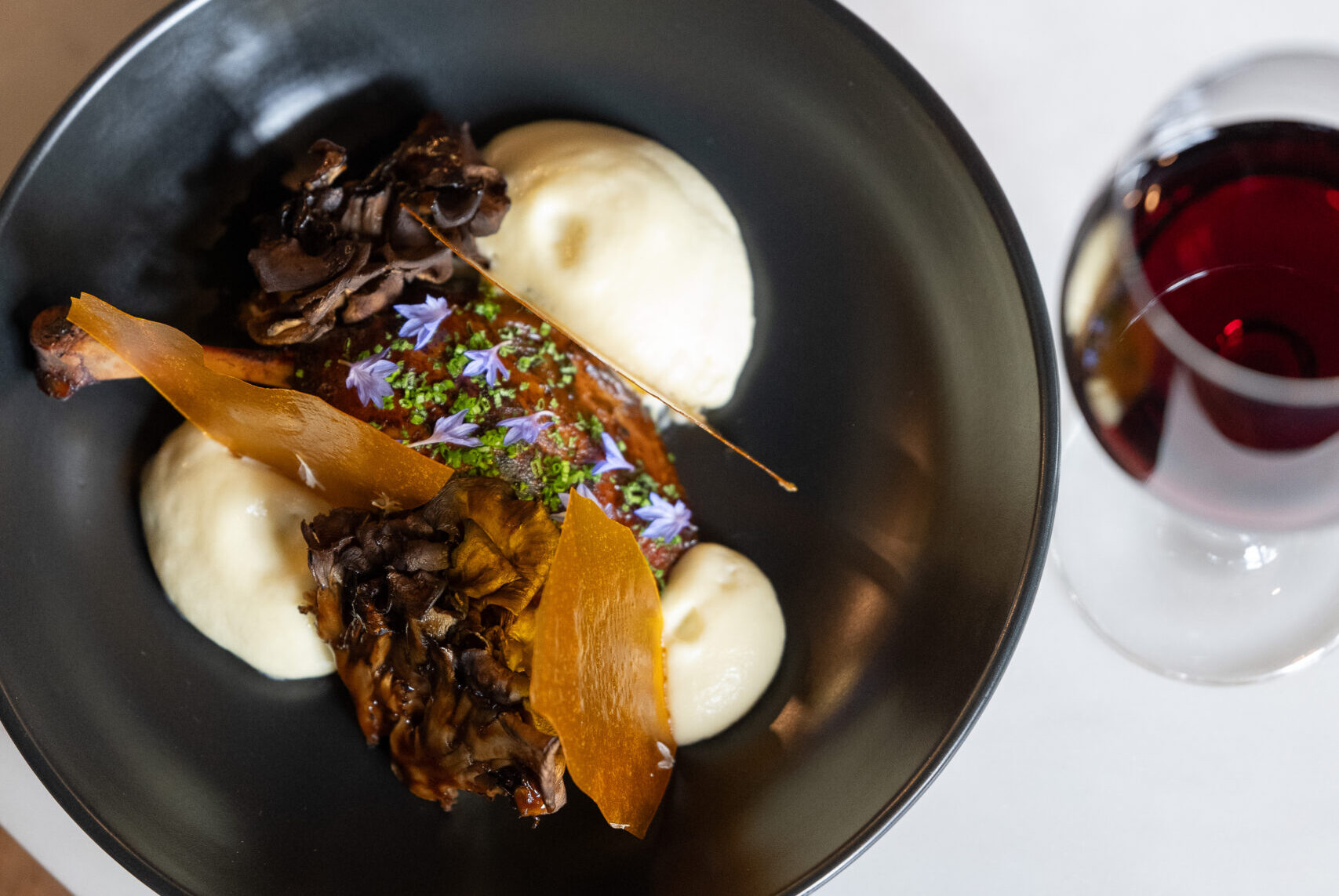 A black bowl contains a serving of mashed potatoes paired with dark mushrooms and garnished with orange crispy elements and small edible flowers. Next to the bowl, there is a glass of red wine resting on a white marble surface.