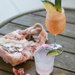 A wooden table displays two cocktails: a tall orange drink garnished with a pineapple and a small pink drink with a lime slice, both adorned with cocktail umbrellas. Nearby is a pink embellished bag and a pair of orange sunglasses.