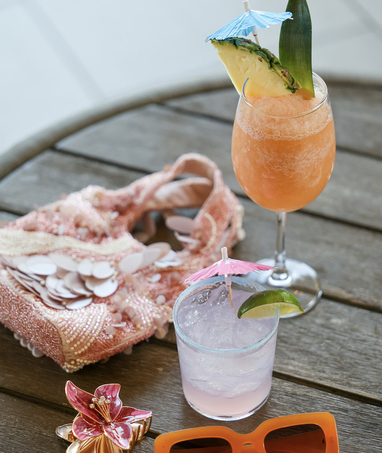 A wooden table displays two cocktails: a tall orange drink garnished with a pineapple and a small pink drink with a lime slice, both adorned with cocktail umbrellas. Nearby is a pink embellished bag and a pair of orange sunglasses.