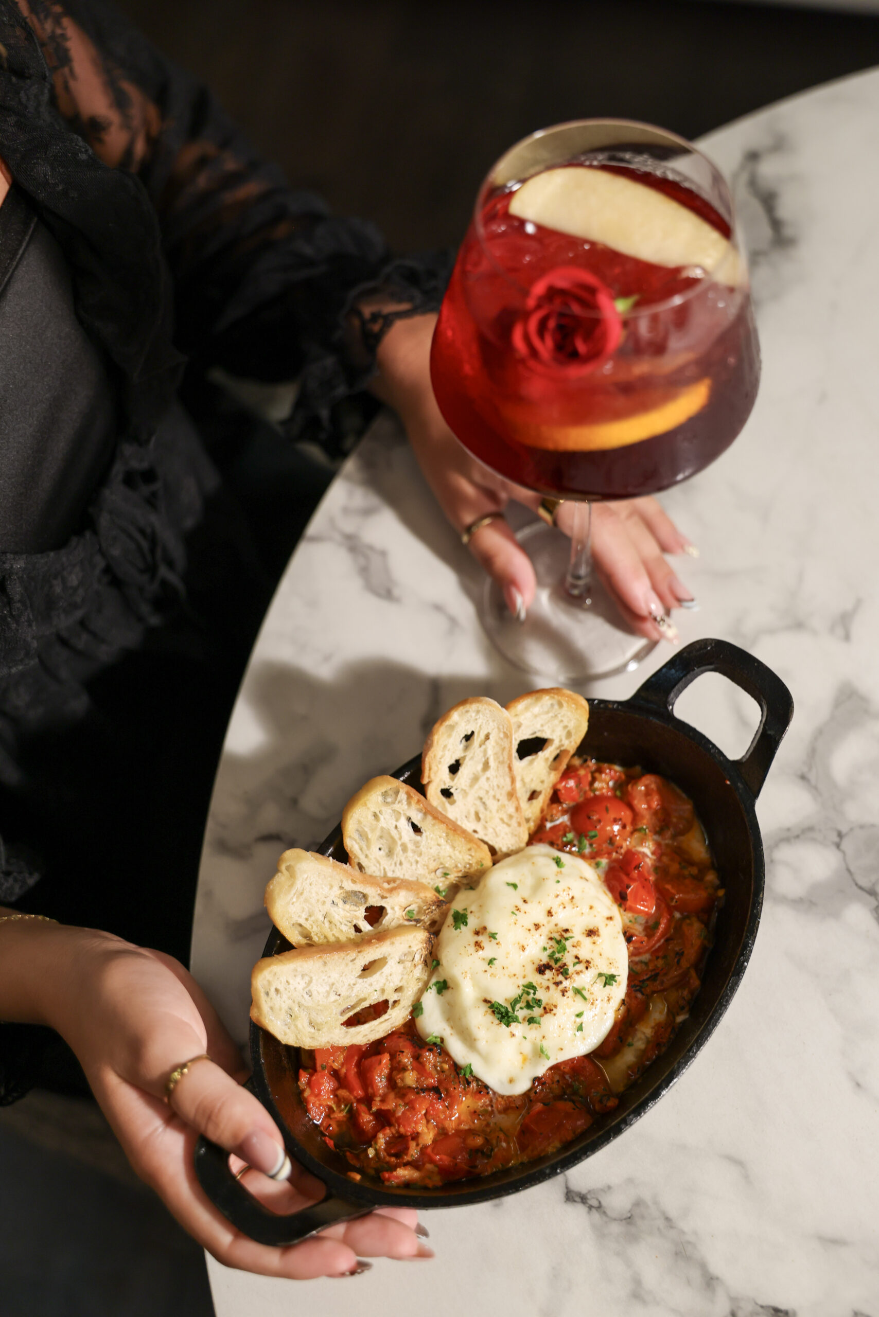 A person holds a black skillet of baked pasta topped with melted cheese and toasted bread slices, with a red cocktail on a marble table.
