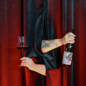 A person in a black draped costume holds a wine glass and a bottle in front of red and black fabric curtains.