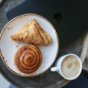 A white plate holds a triangular pastry and a cinnamon roll on a round black table beside a small cup of coffee.