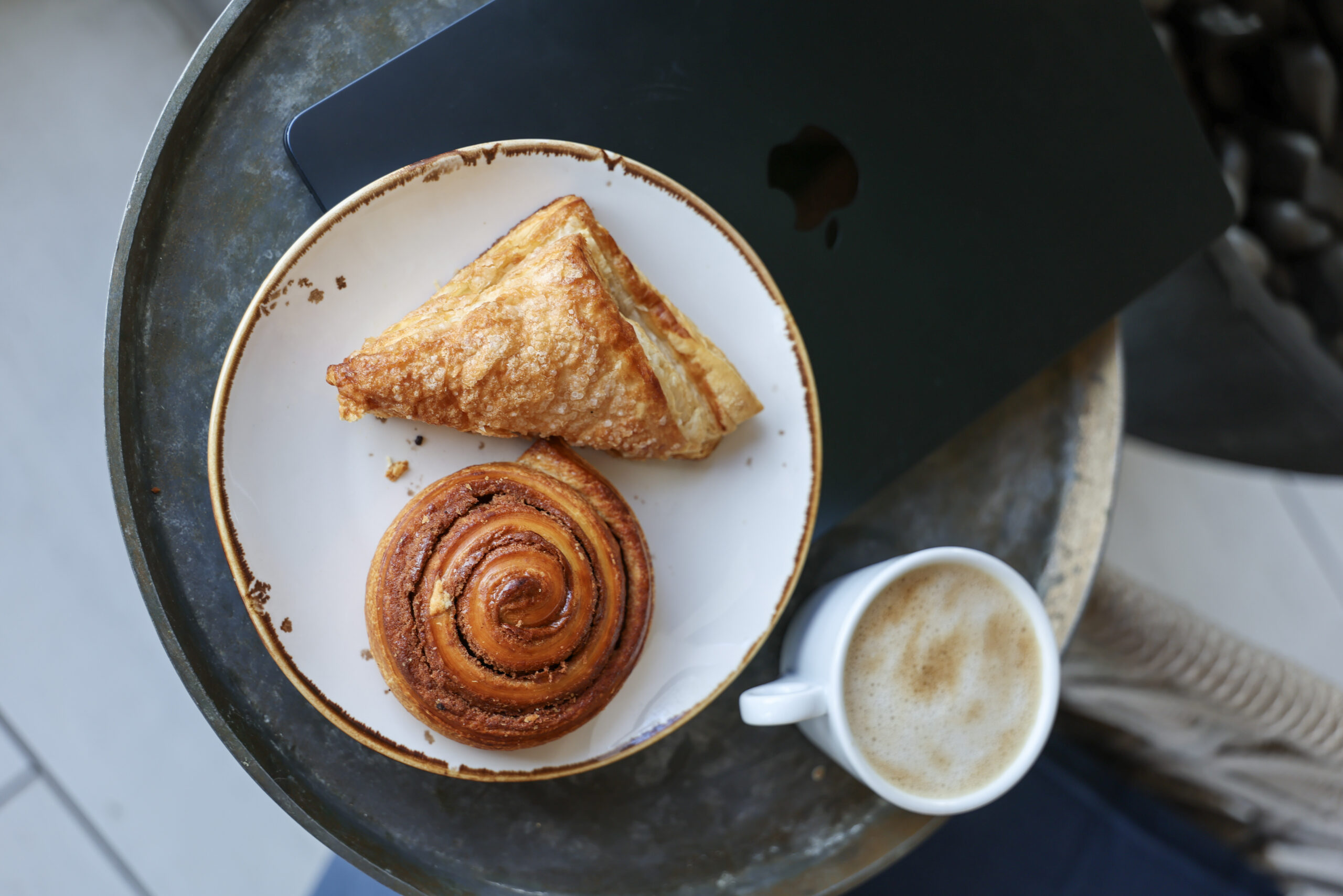 A white plate holds a triangular pastry and a cinnamon roll on a round black table beside a small cup of coffee.