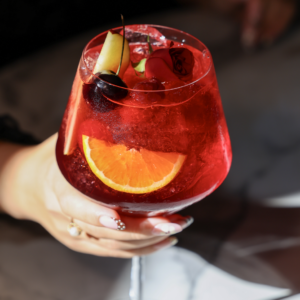 A hand with manicured nails holds a stemmed glass of red cocktail garnished with an orange slice, apple wedge, and rose-shaped decoration against a dark background.