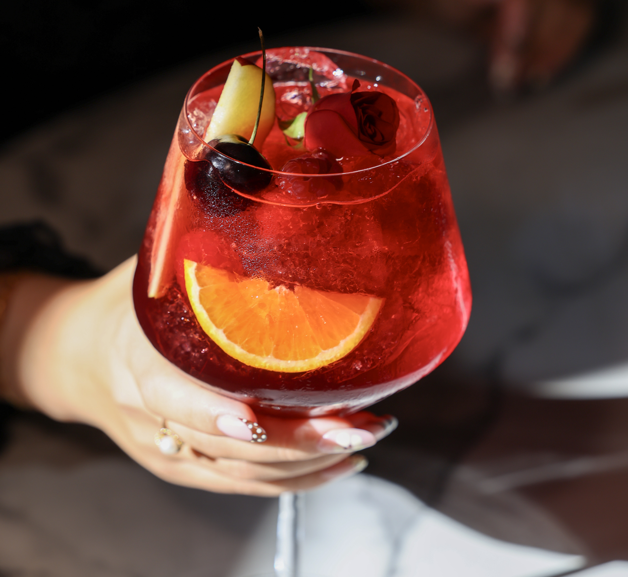 A hand with manicured nails holds a stemmed glass of red cocktail garnished with an orange slice, apple wedge, and rose-shaped decoration against a dark background.