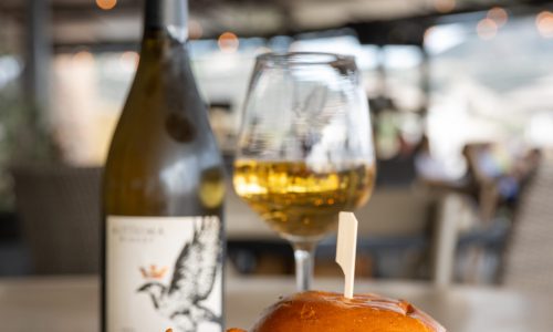 A plate featuring a chicken burger with lettuce, served alongside curly fries. A glass of white wine and a bottle of wine are visible in the background, with a blurred restaurant setting and soft lighting.