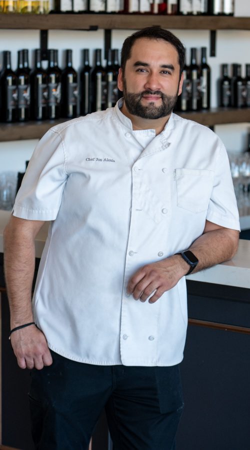 A chef posing confidently at a winery bar, wearing a white chef's coat with his name embroidered. Behind him is a rack of wine bottles, showcasing the winery's selection.