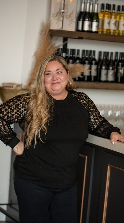 A woman with long hair wearing a black blouse stands at a bar with bottles of wine displayed behind her. She poses confidently with one hand on the counter, representing the Winery Team.